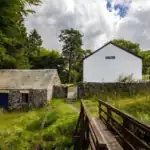Casa rural blanca y edificio de piedra junto a un pequeño puente de madera en una aldea verde del Camino, rodeada de naturaleza y silencio