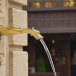 Fuente de agua con caño dorado en forma de cabeza, el agua fluye de manera constante hacia una pila de piedra. Imagen simbólica sobre flujo y equilibrio.