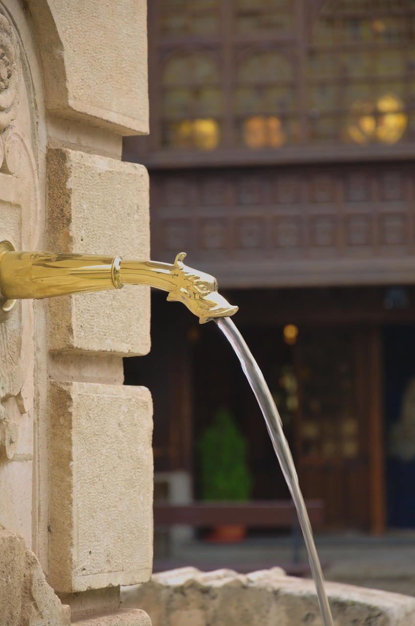 Fuente de agua con caño dorado en forma de cabeza, el agua fluye de manera constante hacia una pila de piedra. Imagen simbólica sobre flujo y equilibrio.